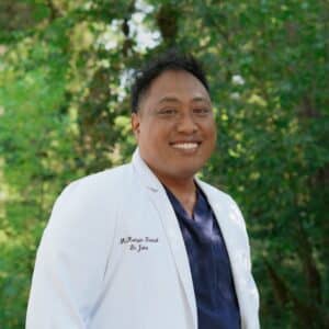 Dr. Ioane Ofisa in whitecoat and scrubs with forest background